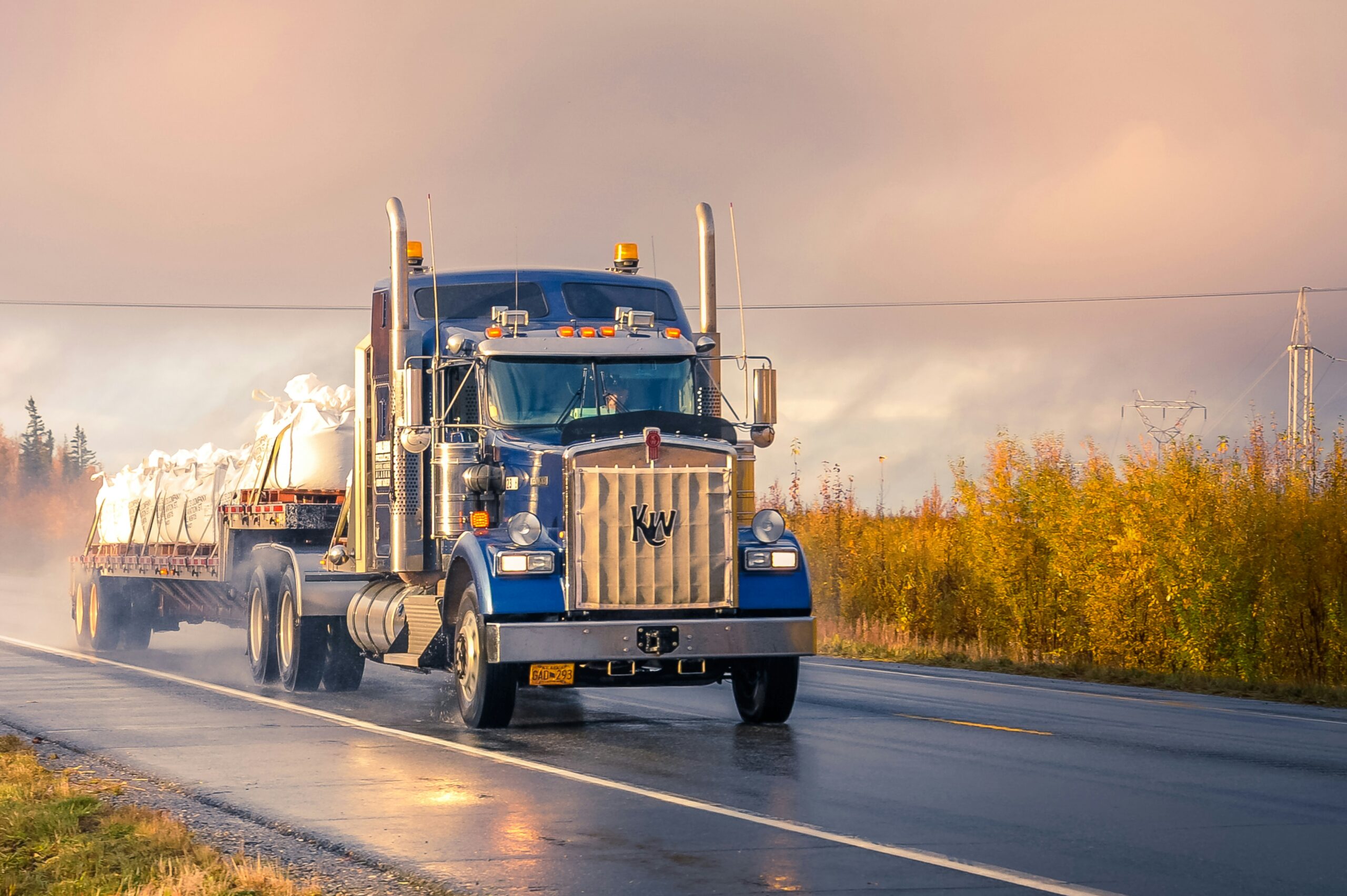 image of a flat bed truck with hay bales driving on the road with the road reflecting the morning sun